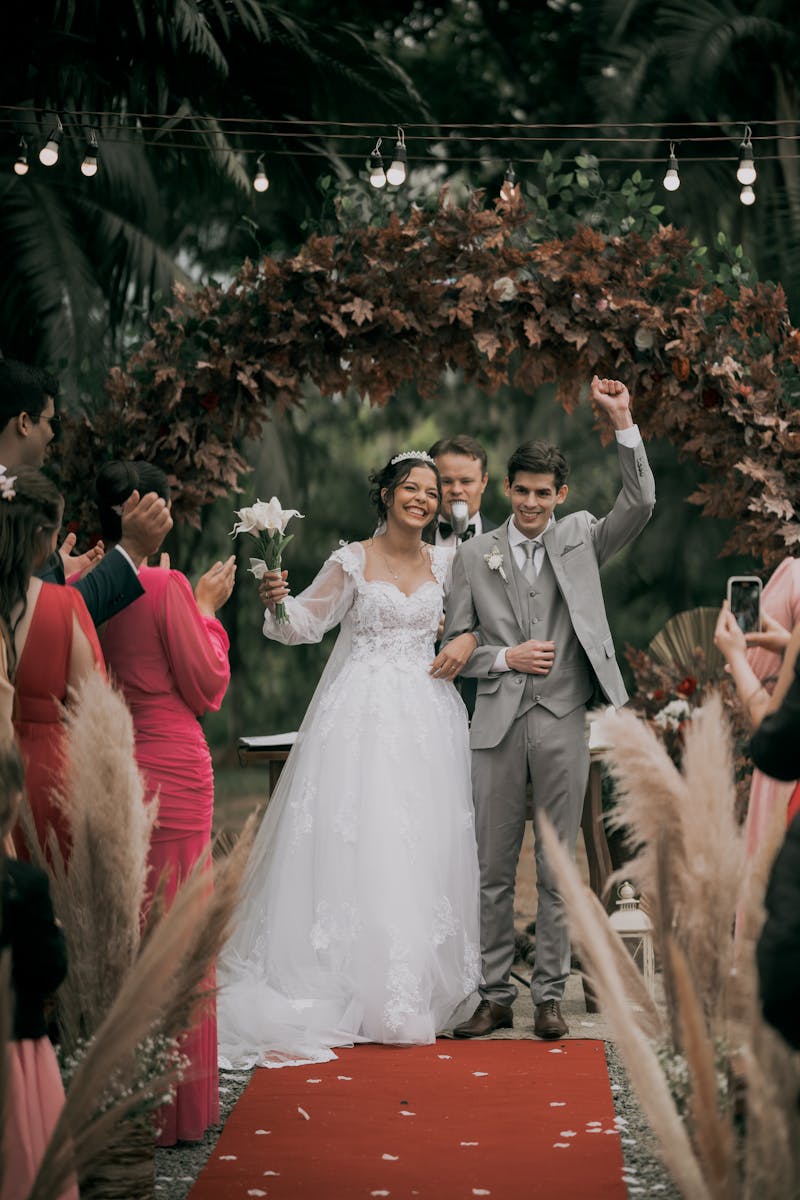 Happy couple celebrates their wedding under a floral arch with loved ones cheering on a festive day.