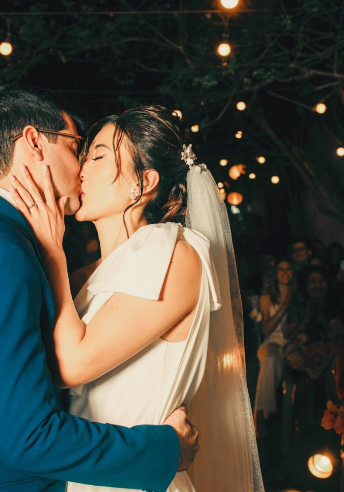 Newlywed couple sharing a romantic kiss under enchanting outdoor string lights.